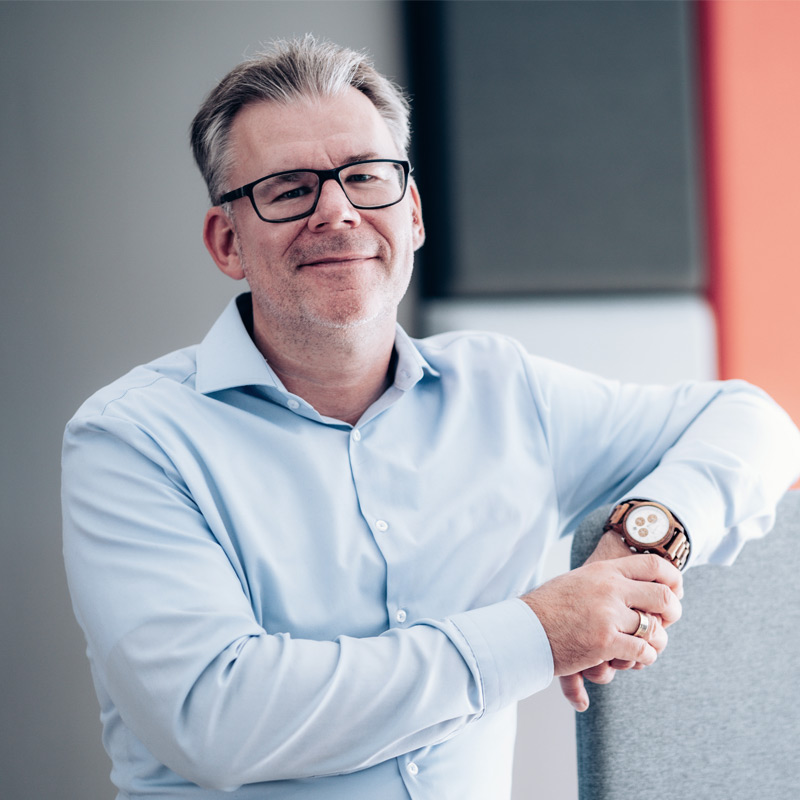 Mann mit hellblauem Hemd und Brille lächelt entspannt.